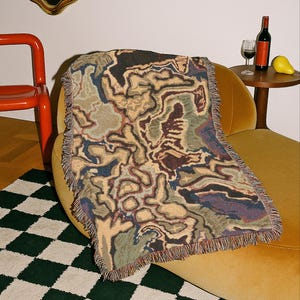 Könnte beinhalten: Eine gewebte Decke mit einem einzigartigen abstrakten Muster in Beige-, Braun-, Blau- und Grüntönen. Die Decke hat einen Fransenrand und ist über einen gelben Stuhl drapiert. Eine Weinflasche und ein Glas stehen auf einem Beistelltisch.