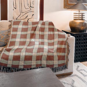 May include: A woven throw blanket with a plaid pattern in shades of red, green, and beige, draped over a beige sofa. The blanket has a fringed edge. The scene includes a lamp and a decorative side table.