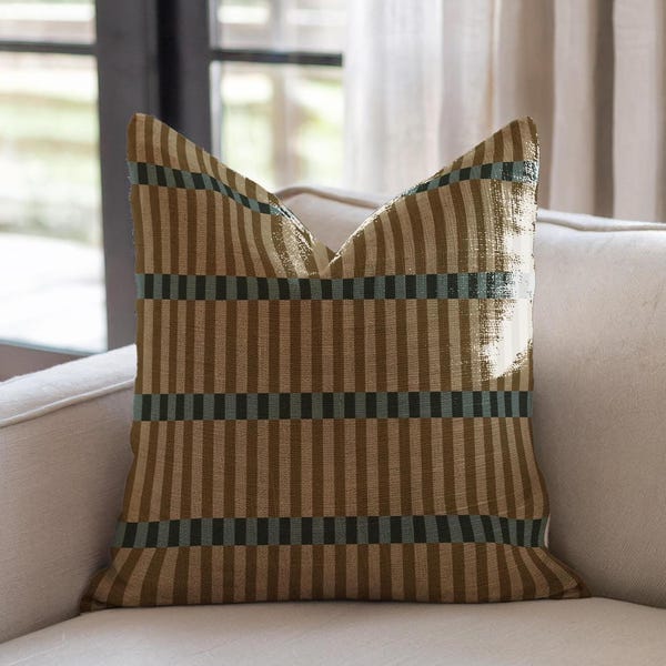 Pode incluir: Almofada decorativa quadrada com um padrão listrado em tons de castanho, verde oliva e verde-azulado escuro. A almofada está colocada em um sofá creme, com uma janela e cortinas ao fundo.