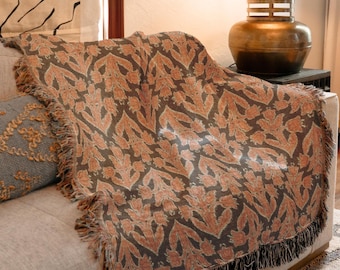 Op de afbeelding: Een decoratieve sprei met een bloemenpatroon in bruin- en oranjetinten, gedrapeerd over een beige bank. De sprei heeft een gefranjerde rand in zwart. Een grijs kussen met gouden borduursel is zichtbaar.