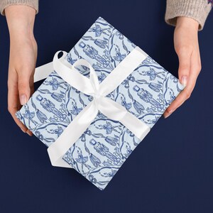 May include: A wrapped gift with a white ribbon, held by two hands. The gift wrap is light blue with a repeating pattern of blue nutcrackers and ballerinas. The background is a solid dark blue.