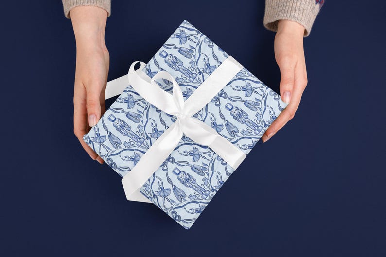 May include: A wrapped gift with a white ribbon, held by two hands. The gift wrap is light blue with a repeating pattern of blue nutcrackers and ballerinas. The background is a solid dark blue.