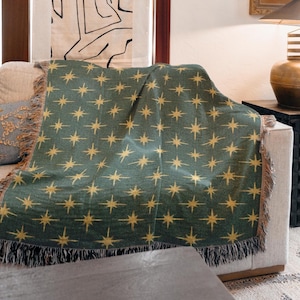 May include: A green throw blanket with a repeating gold starburst pattern. The blanket is draped over a beige sofa and has a fringed edge. The throw is a decorative home textile.