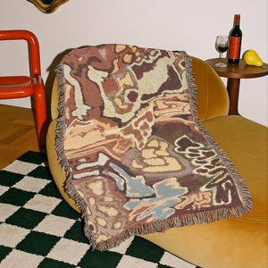 May include: A woven throw blanket with a brown, beige, and blue abstract pattern, fringed edges, draped over a yellow chair. A green and white checkered rug is in the foreground. A small table holds a wine bottle, glass, and a pear.