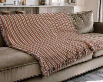 Puede incluir: Una manta a rayas rojas y verdes con flecos, colocada sobre un sofá beige. La manta presenta rayas verticales en diferentes tonos de rojo, verde y crema, creando una estética acogedora. Los flecos añaden un toque de textura.