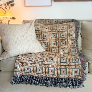 Könnte beinhalten: Beige Überwurfdecke mit geometrischem Muster in Braun, Grün und Rot, drapiert über einem Sofa. Die Decke hat einen Fransenrand in Blau-, Grün- und Rottönen. Ein beiges Kissen liegt neben der Decke.