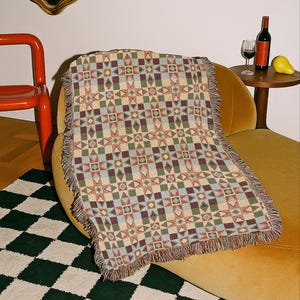 May include: A woven throw blanket featuring a repeating geometric pattern in muted shades of burgundy, sage green, light blue, and cream. The rectangular blanket has a fringed edge along the sides and displays a complex grid of stars and squares.