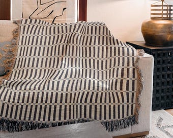 May include: A beige and dark gray patterned throw blanket draped over a sofa. The blanket features a repeating vertical stripe design with a fringed edge. The sofa is neutral-toned, and the background includes a lamp and artwork.
