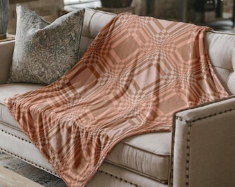 May include: A peach-colored blanket with a plaid pattern draped over a beige sofa. The blanket features a repeating design of squares and lines in varying shades of peach and brown. A decorative pillow rests on the sofa.