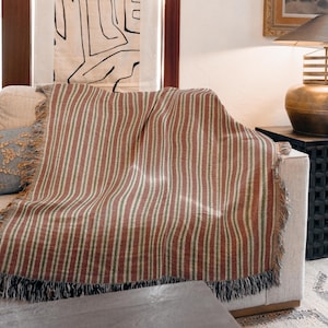 Peut inclure: Une couverture à rayures rouges, vertes et crème est drapée sur un canapé beige. La couverture a une bordure à franges et un motif à rayures verticales. Le canapé est dans un cadre de salon.