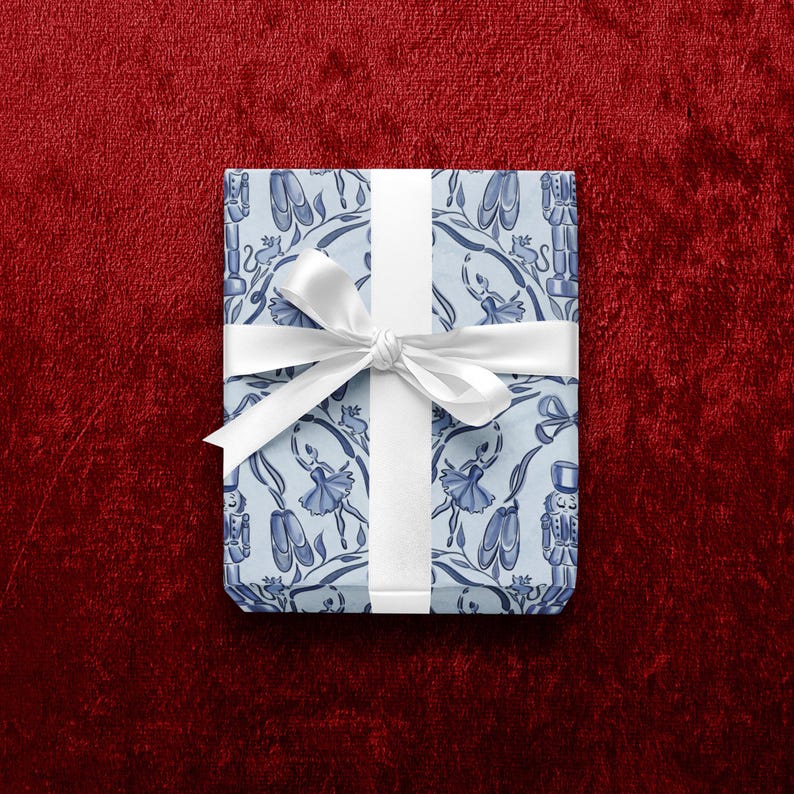 May include: A wrapped gift with light blue wrapping paper featuring a pattern of ballet dancers, pointe shoes, and floral elements. The gift is tied with a white satin ribbon and bow. The background is a deep red velvet.