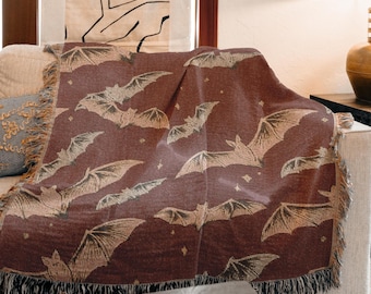 May include: A burgundy throw blanket with a repeating pattern of bats in a beige color. The blanket has a fringed edge and is draped over a sofa. The bats are depicted with their wings spread, and there are small star-like shapes scattered throughout.