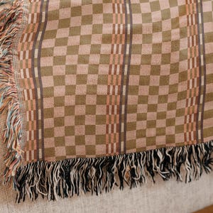May include: A woven throw blanket with a checkered pattern in shades of brown and tan. The blanket has vertical stripes and fringed edges in black, tan, and other colours. The blanket is resting on a light-coloured surface.
