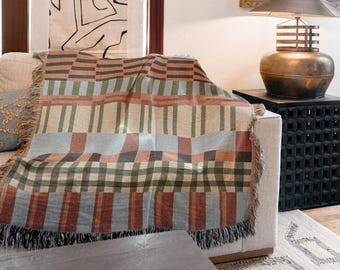 Puede incluir: Una manta tejida con un patrón geométrico multicolor con rayas horizontales y verticales en tonos verde salvia, naranja terracota, azul claro y crema, terminada con un borde de flecos y colocada sobre un sofá.