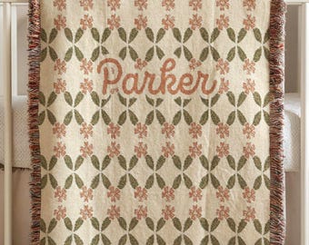 May include: Cream-colored baby blanket with a floral pattern in muted red and green. The name "Parker" is embroidered in a matching red hue. The blanket has a fringed edge and is draped over a crib.