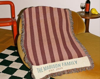 May include: A striped throw blanket in shades of burgundy and tan, draped over a yellow armchair. The blanket has a fringed edge and the text "THE MADISON FAMILY EST. 2025" in a light blue color. A bottle of wine and a pear are on a side table.
