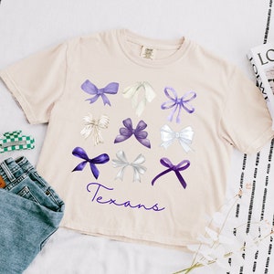 May include: A light beige t-shirt with a graphic of purple and white bows and the word "Texans" printed on the front.