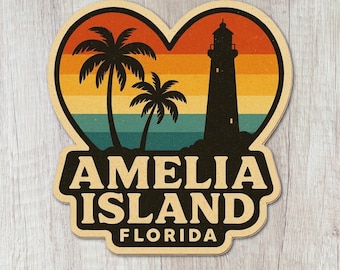 May include: A heart-shaped sticker featuring a sunset gradient background with two black palm tree silhouettes and a lighthouse. The text AMELIA ISLAND FLORIDA is printed in bold cream-colored letters on a dark brown base at the bottom of the sticker.