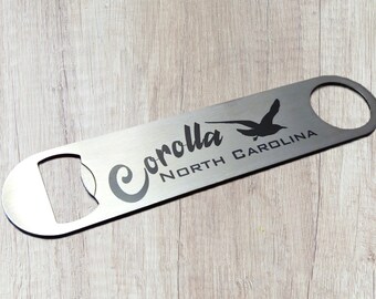 May include: A flat, silver-toned metal bottle opener with a rounded handle featuring a circular hole at one end. The surface is engraved with a black graphic of a flying bird and the text "Corolla NORTH CAROLINA" in a mix of script and block fonts.