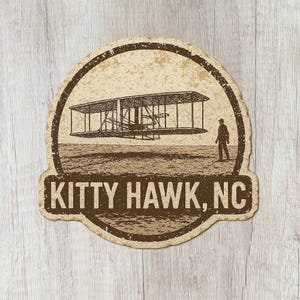 May include: A vintage-style sticker featuring a sepia-toned illustration of the Wright brothers' plane in flight over a field, with a person standing nearby. The text "KITTY HAWK, NC" is displayed below the image.