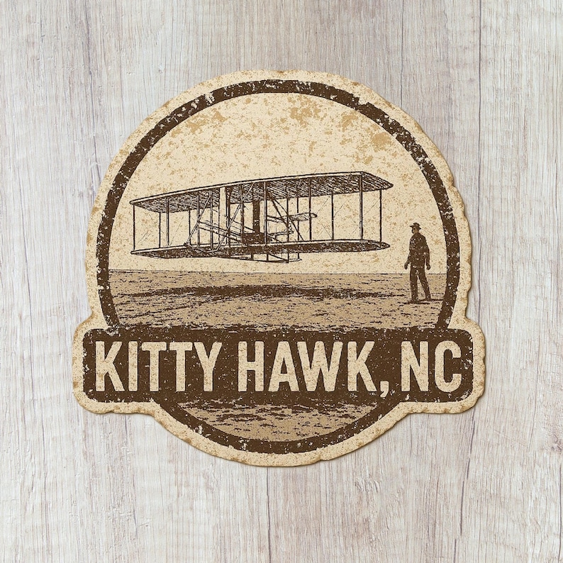 May include: A vintage-style sticker featuring a sepia-toned illustration of the Wright brothers' plane in flight over a field, with a person standing nearby. The text "KITTY HAWK, NC" is displayed below the image.