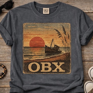 May include: Dark gray t-shirt featuring a distressed graphic of a sunset over the ocean with a beached boat. The graphic includes the text "OBX" in a vintage style. The shirt is made of a soft material and has the "Comfort Colors" label.