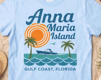 May include: A light blue short-sleeve t-shirt featuring a graphic print with the text Anna Maria Island and GULF COAST, FLORIDA. The design includes an orange sun, two green palm trees, a blue boat, and stylized blue ocean waves.