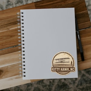 May include: A spiral-bound notebook with a sticker featuring an airplane design and the text "KITTY HAWK, NC." A black pen rests on the right side of the notebook, which is placed on a wooden surface.
