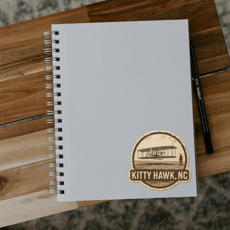 May include: A spiral-bound notebook with a sticker featuring an airplane design and the text "KITTY HAWK, NC." A black pen rests on the right side of the notebook, which is placed on a wooden surface.