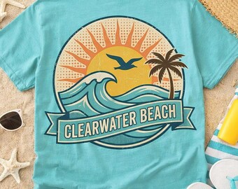 May include: A turquoise t-shirt featuring a circular graphic on the back. The design displays a yellow sun, a blue ocean wave, a palm tree, and a flying bird. A banner across the bottom contains the text CLEARWATER BEACH in bold letters.