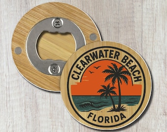 May include: A circular bamboo bottle opener featuring a graphic of a sunset over the ocean with palm trees and birds. The text reads Clearwater Beach Florida. The metal opener mechanism is visible in the center of the bamboo wood frame.