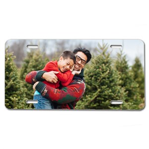 May include: A father and son are smiling and hugging in a Christmas tree lot. The father is wearing a red and green sweater and the son is wearing a red sweater.