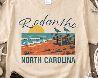 May include: A sand-colored short-sleeve t-shirt featuring a graphic print of a beach scene with a yellow sun, blue ocean, orange sand, and a wooden pier with two birds. The text reads Rodanthe and NORTH CAROLINA in dark teal lettering.