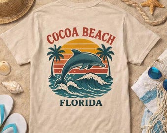 May include: A beige short-sleeve t-shirt featuring a graphic print of a dolphin jumping over ocean waves with palm trees and a sunset. The text COCOA BEACH is arched above the sun, and the word FLORIDA is printed in dark blue below the waves.