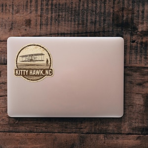 May include: A sticker featuring a sepia-toned illustration of the Wright brothers' plane at Kitty Hawk, NC. The design is circular with the text "KITTY HAWK, NC" at the bottom. The sticker is placed on a laptop against a wooden background.
