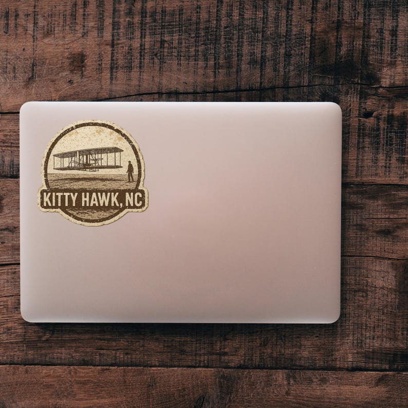May include: A sticker featuring a sepia-toned illustration of the Wright brothers' plane at Kitty Hawk, NC. The design is circular with the text "KITTY HAWK, NC" at the bottom. The sticker is placed on a laptop against a wooden background.