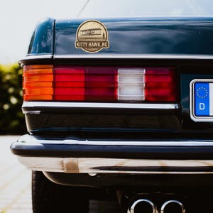 May include: A dark green car's rear section with a sticker featuring an illustration of an early airplane and the text "KITTY HAWK, NC". The car's tail lights are red and orange, with a chrome bumper and a black tire.