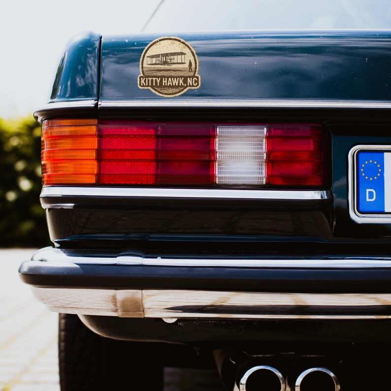 May include: A dark green car's rear section with a sticker featuring an illustration of an early airplane and the text "KITTY HAWK, NC". The car's tail lights are red and orange, with a chrome bumper and a black tire.
