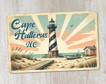 May include: A rectangular print featuring a black and white striped lighthouse, a wooden boardwalk, a small boat, and a cabin on a sandy beach. The background displays a sunburst pattern in soft blue and peach tones with the text Cape Hatteras NC.