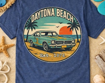 May include: A heather blue t-shirt featuring a circular graphic on the back. The design depicts a teal vintage car with the number 3 on the side, parked on a beach with palm trees, an orange sun, and the text Daytona Beach Est. 1876.