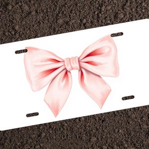 May include: A white license plate with a pink bow in the center. The bow is tied with a large knot and the ends of the ribbon are flowing.