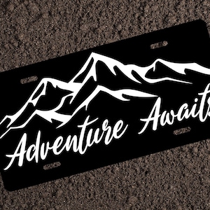 May include: Black and white mountain range design on a black license plate with the text "Adventure Awaits".