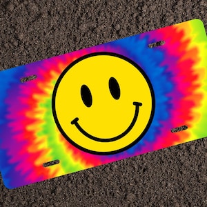 May include: A yellow smiley face on a tie-dye license plate. The license plate is a bright rainbow of colors, including pink, blue, green, and yellow.