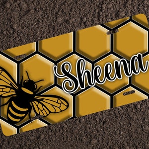 May include: A license plate featuring a bee and honeycomb design. The plate has a yellow and black honeycomb pattern with a black and yellow bee on the left. The name "Sheena" is written in a cursive font.