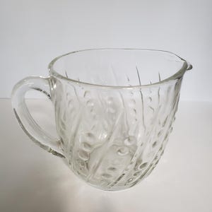 May include: Clear glass pitcher with a handle and a spout. The pitcher has a textured design of vertical lines and small circles. The pitcher is empty and sits on a white surface.