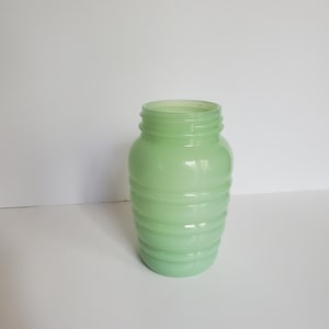 May include: A light green glass jar with a ribbed design. The jar has a wide opening and a rounded body. The glass has a translucent quality, and the jar is sitting on a white surface.