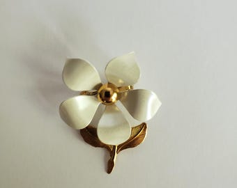 May include: A flower-shaped brooch with five white petals and a gold-colored center. The brooch has a gold-colored stem and leaf design. The brooch is a vintage accessory.
