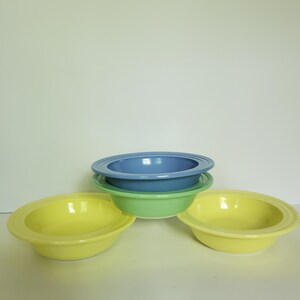 May include: A set of three ceramic bowls with wide rims and concentric circular grooves. The stack includes one light blue bowl positioned on top of a light green bowl, with two bright yellow bowls placed on either side of the base.