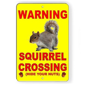 May include: A yellow metal sign with the words "WARNING" and "SQUIRREL CROSSING" in red. A gray squirrel is in the center. The text "(HIDE YOUR NUTS)" is below the crossing text. Two acorn graphics are at the bottom.