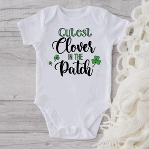May include: White baby onesie with black text that reads "Cutest Clover in the Patch". Green shamrock designs are on the onesie.
